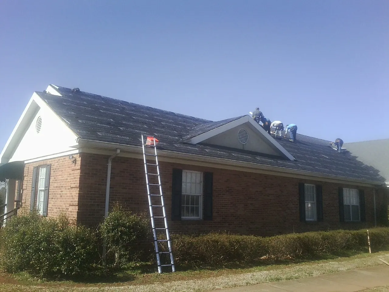 Crew tearing shingles off a brick ranch roof with a ladder set — Metal Roof Repair work by our crew in Perry
